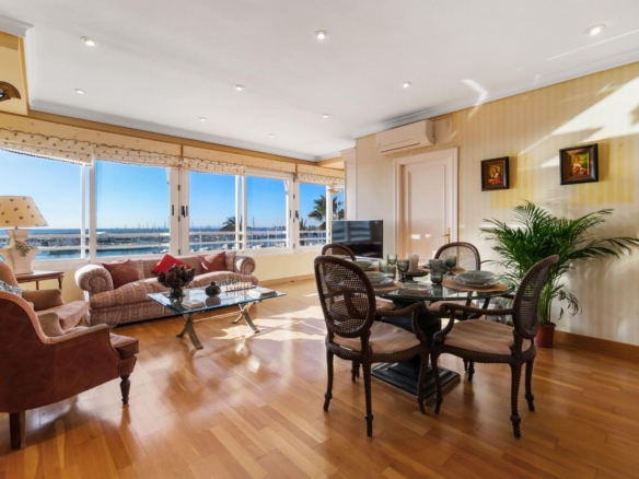 Bright living room with large windows overlooking a marina; pink sofas, glass coffee table, and a glass dining table with woven chairs, wooden floor, and houseplants.