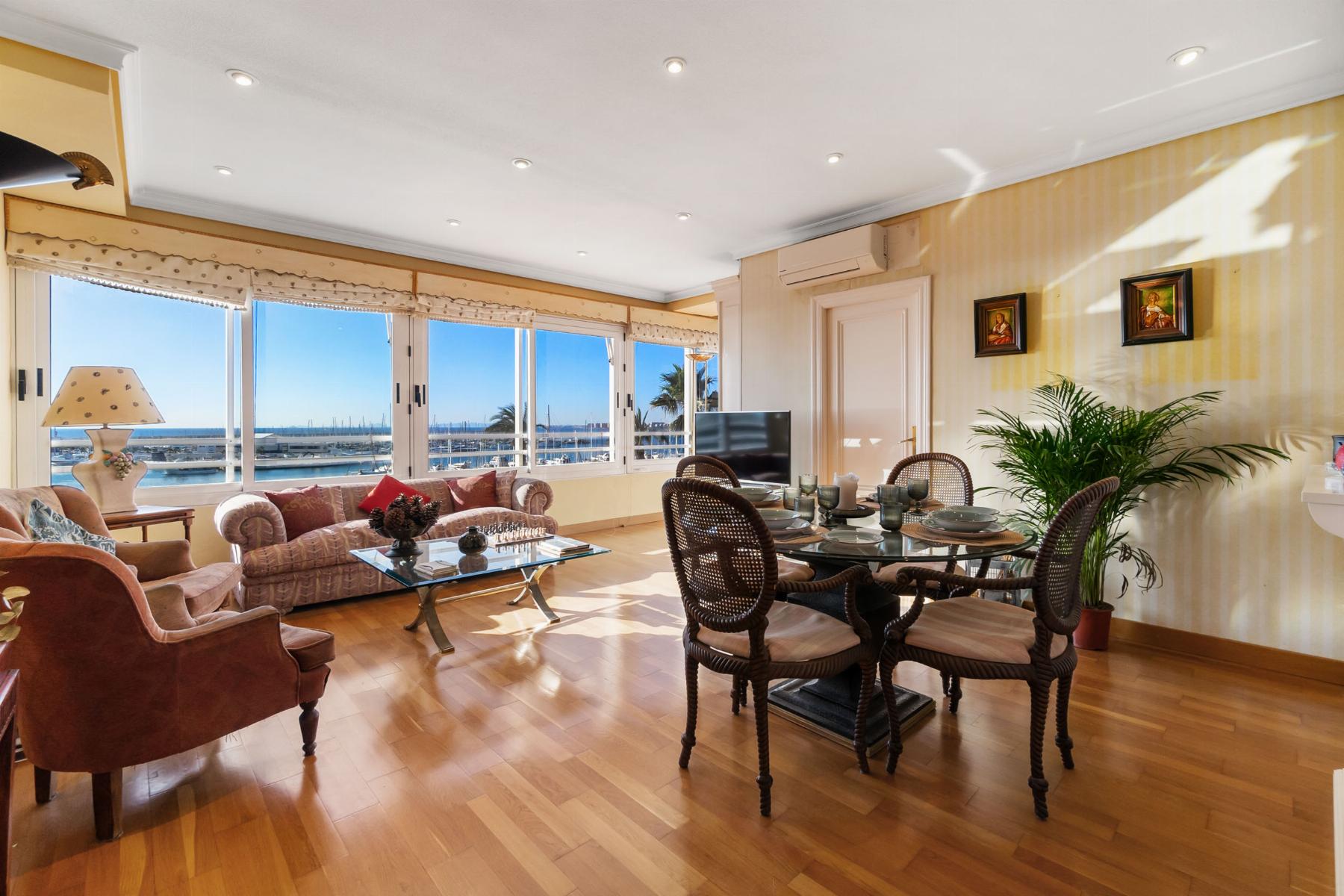 Bright living room with large windows overlooking a marina; pink sofas, glass coffee table, and a glass dining table with woven chairs, wooden floor, and houseplants.