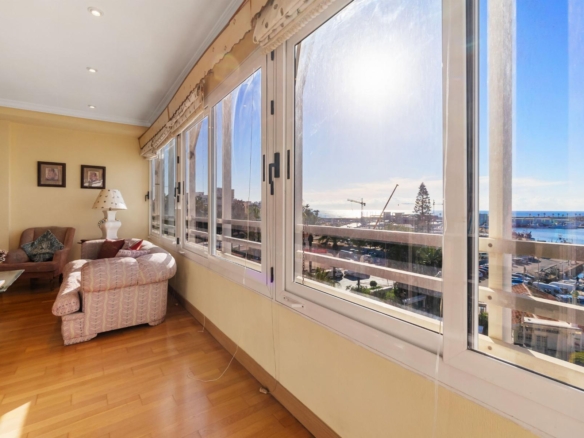 Sunlit living room with pastel pink sofa and armchair facing large windows overlooking a coastal town and sea.