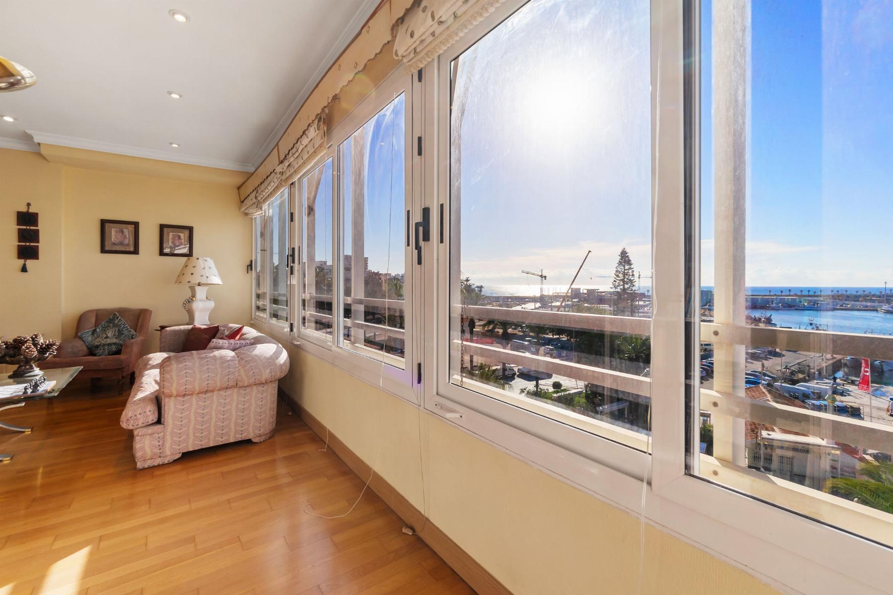 Sunlit living room with pastel pink sofa and armchair facing large windows overlooking a coastal town and sea.