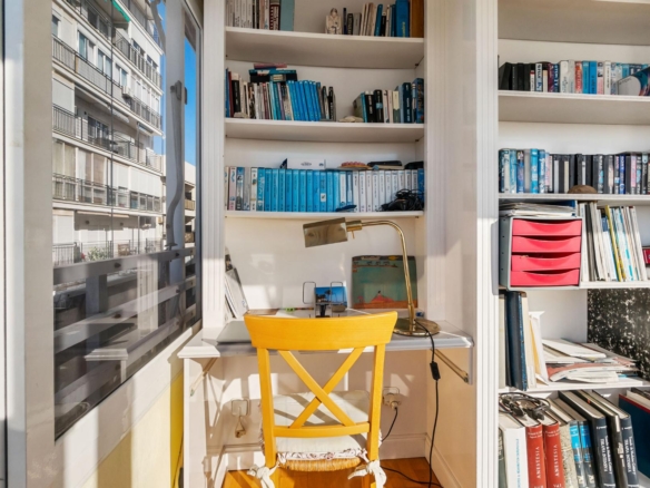 Bright study nook with white built-in shelves filled with blue-toned books, a brass desk lamp, and a yellow chair at a small white desk by a window showing neighboring buildings.