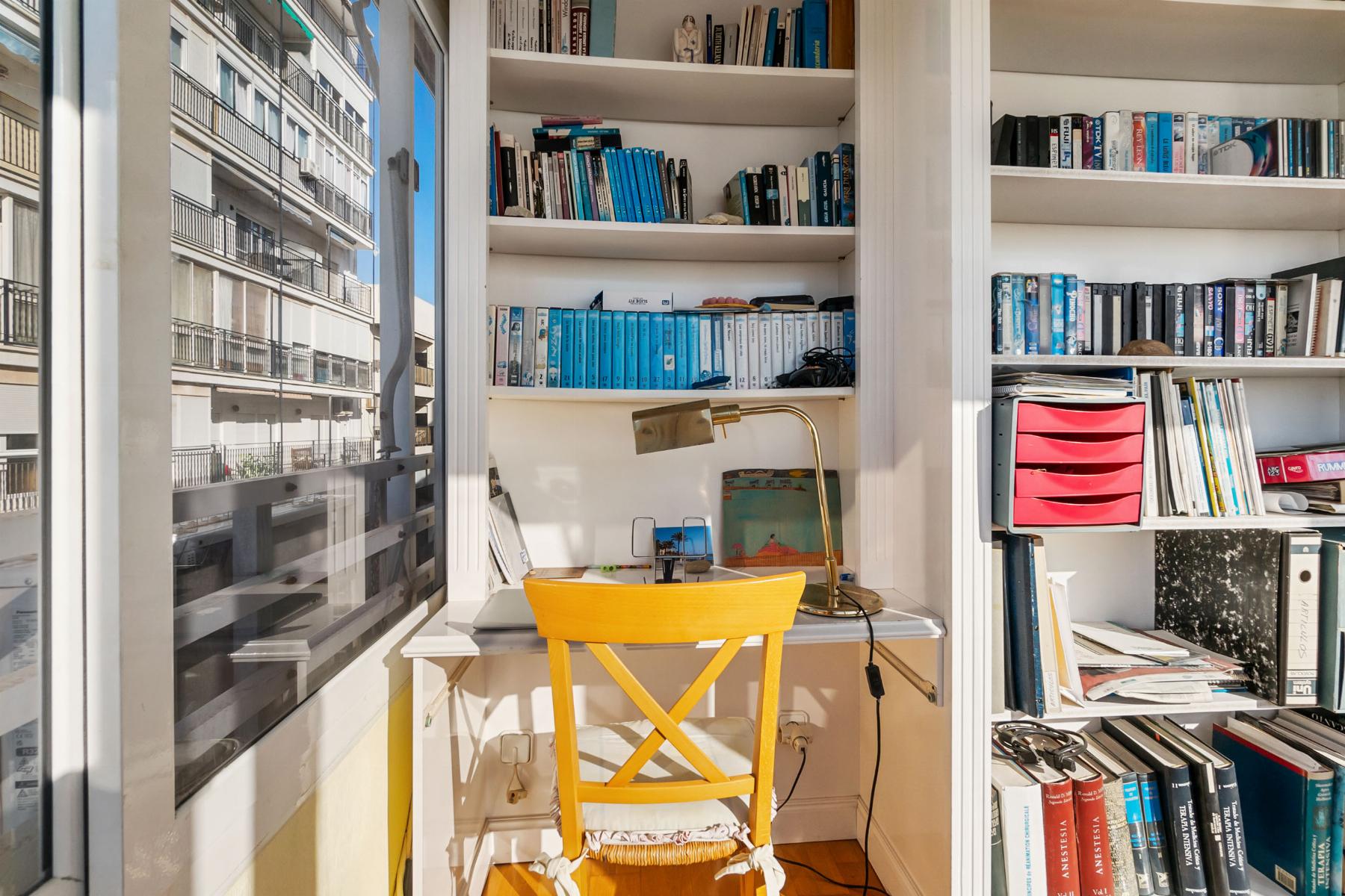 Bright study nook with white built-in shelves filled with blue-toned books, a brass desk lamp, and a yellow chair at a small white desk by a window showing neighboring buildings.