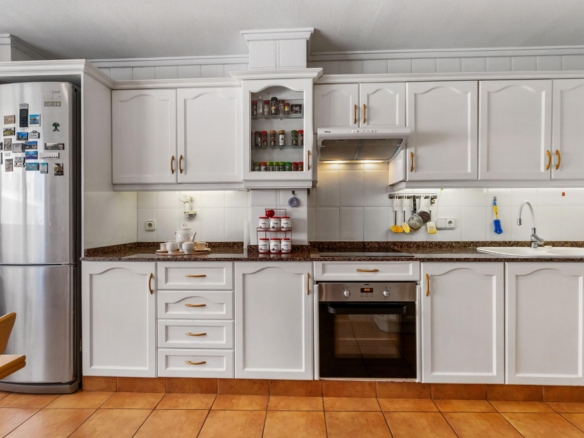 Bright white kitchen with a tall stainless refrigerator on the left, open spice cabinet, and granite countertops.