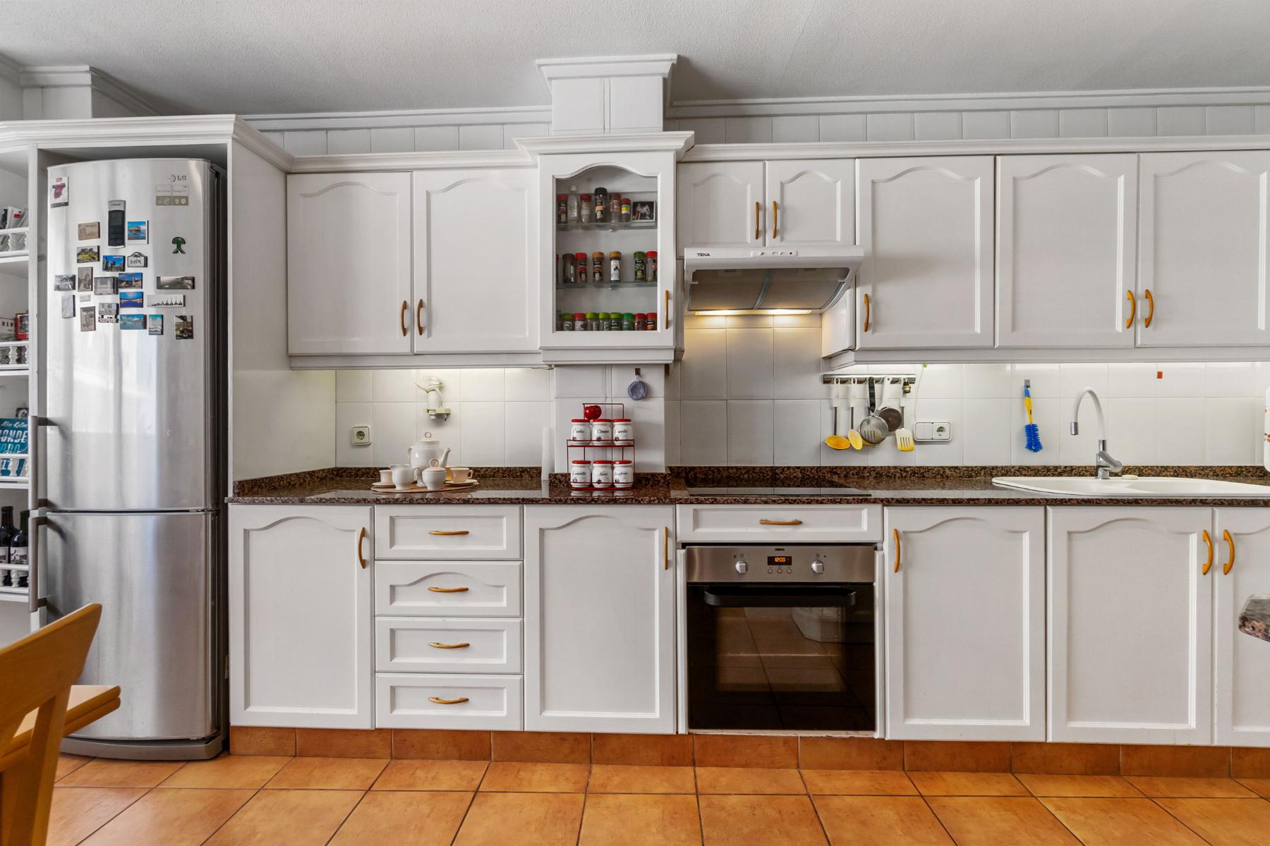 Bright white kitchen with a tall stainless refrigerator on the left, open spice cabinet, and granite countertops.