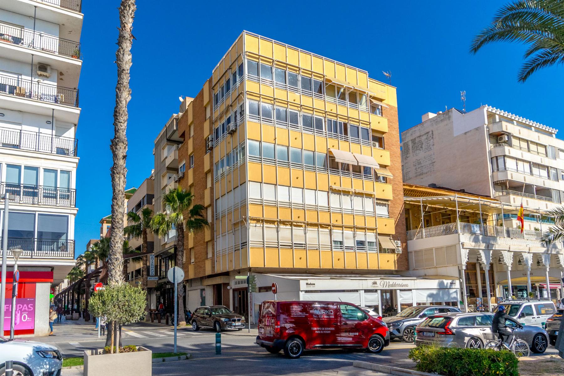 Street scene with a yellow multi-story building, palm trees, blue sky, and parked cars along a storefront-lined street.