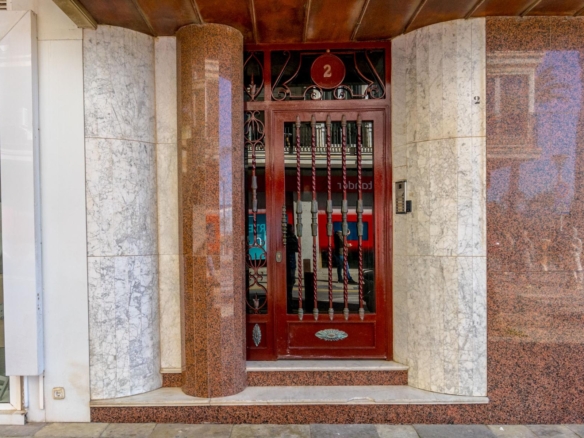 Red ornate metal door with vertical bars set in a marble-tiled storefront façade labeled '2' on a circular plaque above the door.