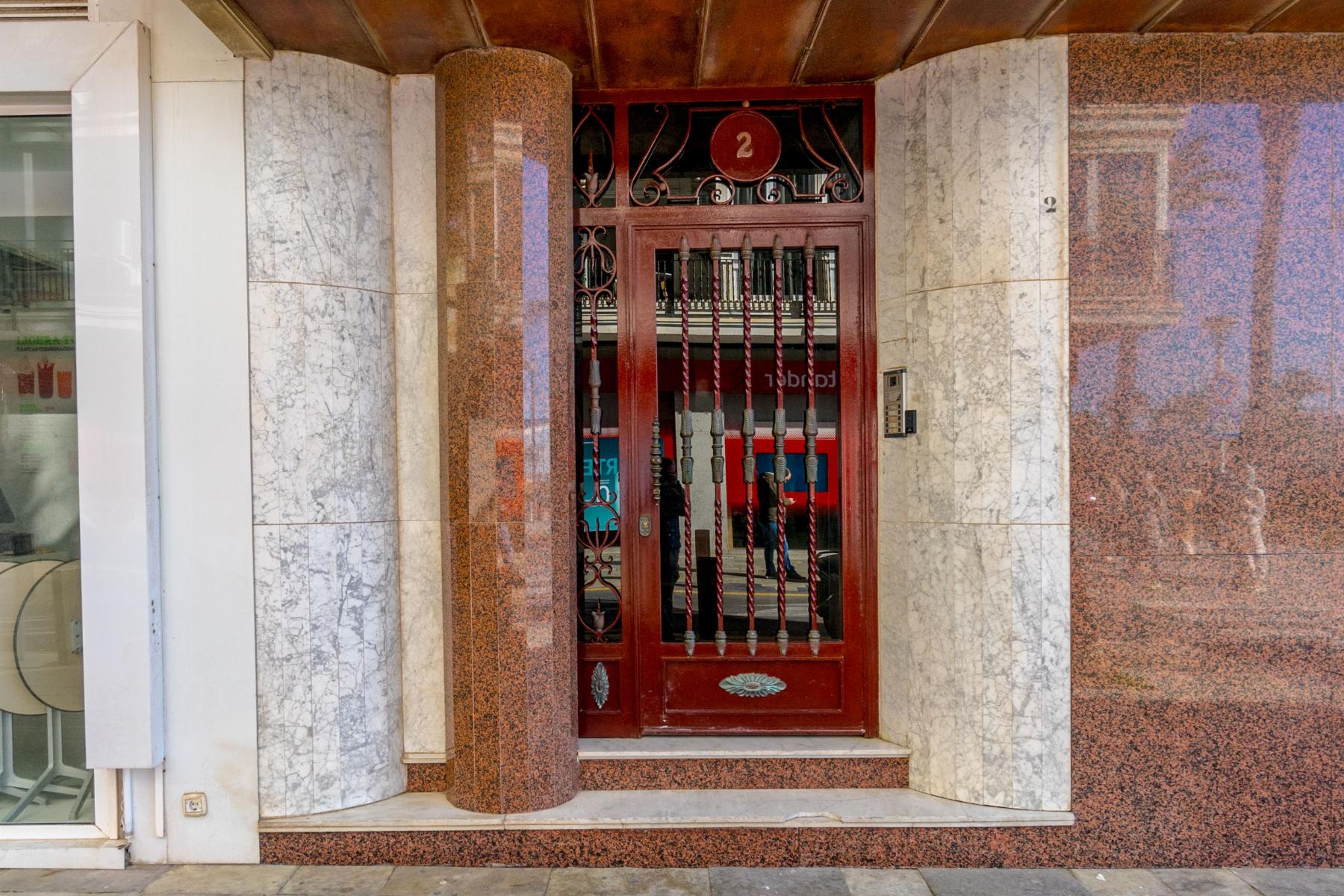Red ornate metal door with vertical bars set in a marble-tiled storefront façade labeled '2' on a circular plaque above the door.