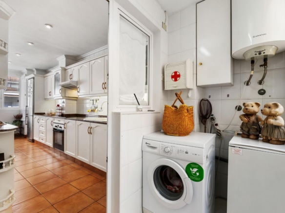 Bright white kitchen with a laundry area: washing machine, wicker basket, and teddy bears on a counter by a water heater; white cabinets and tiled walls dominate the scene.