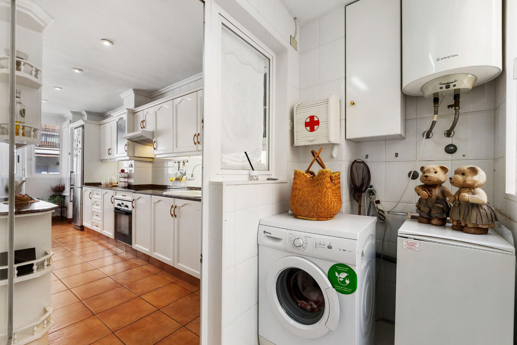 Bright white kitchen with a laundry area: washing machine, wicker basket, and teddy bears on a counter by a water heater; white cabinets and tiled walls dominate the scene.