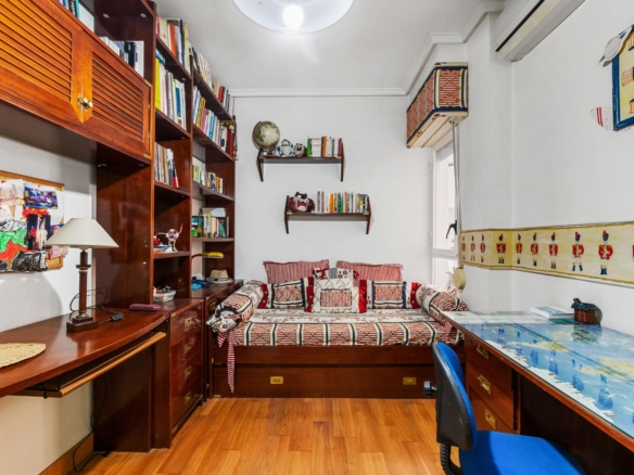 Cozy study with tall wooden bookshelves along left wall, a desk with lamp and photos, and a daybed in the center topped with red‑and‑white cushions; decorative world globe and shelves above add color.