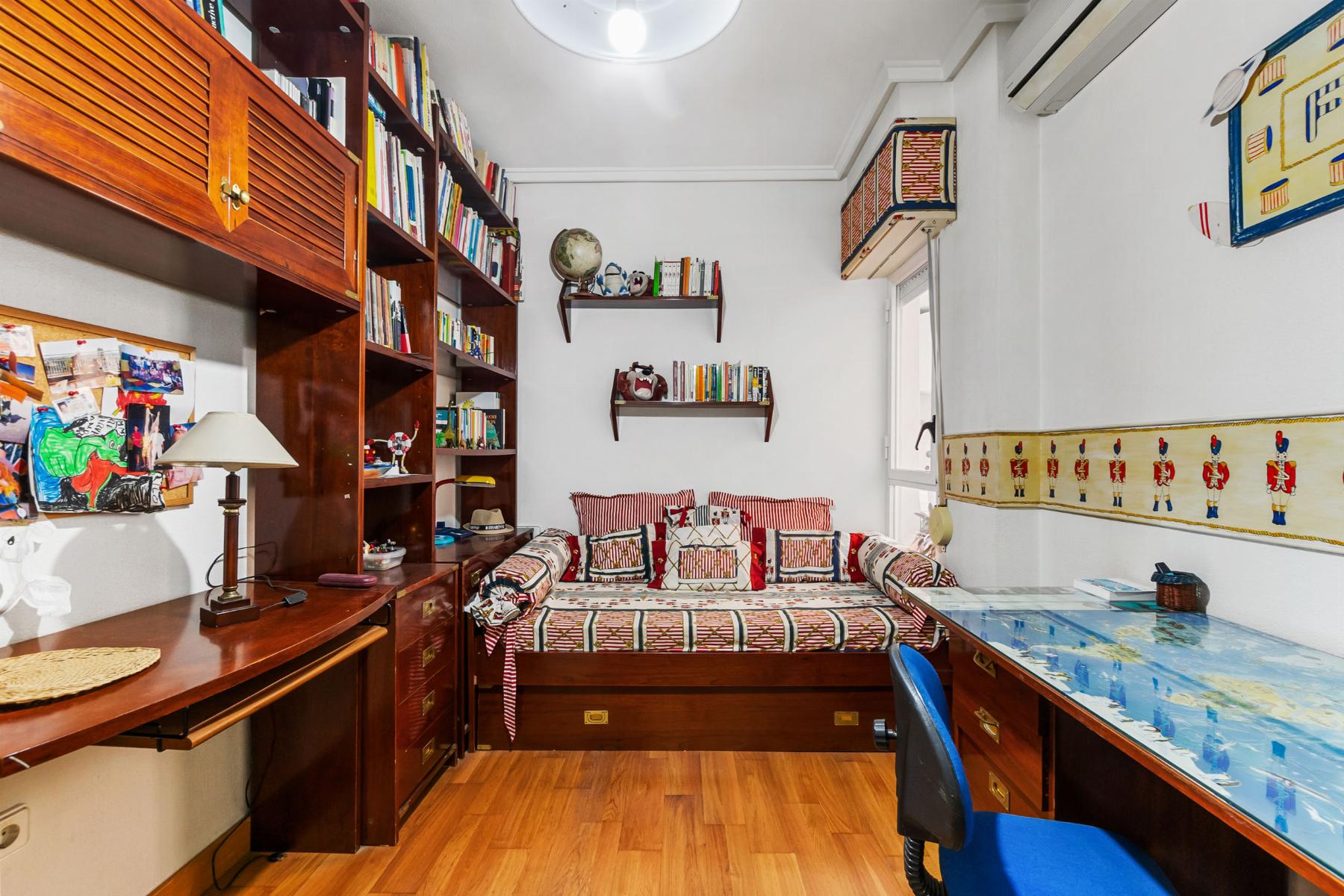 Cozy study with tall wooden bookshelves along left wall, a desk with lamp and photos, and a daybed in the center topped with red‑and‑white cushions; decorative world globe and shelves above add color.