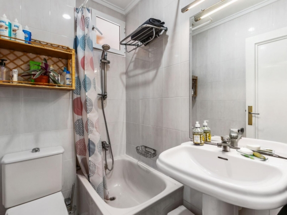 Small white-tiled bathroom with toilet, bathtub/shower combo, colorful patterned curtain, wall shelf of toiletries, and sink with pump bottles.