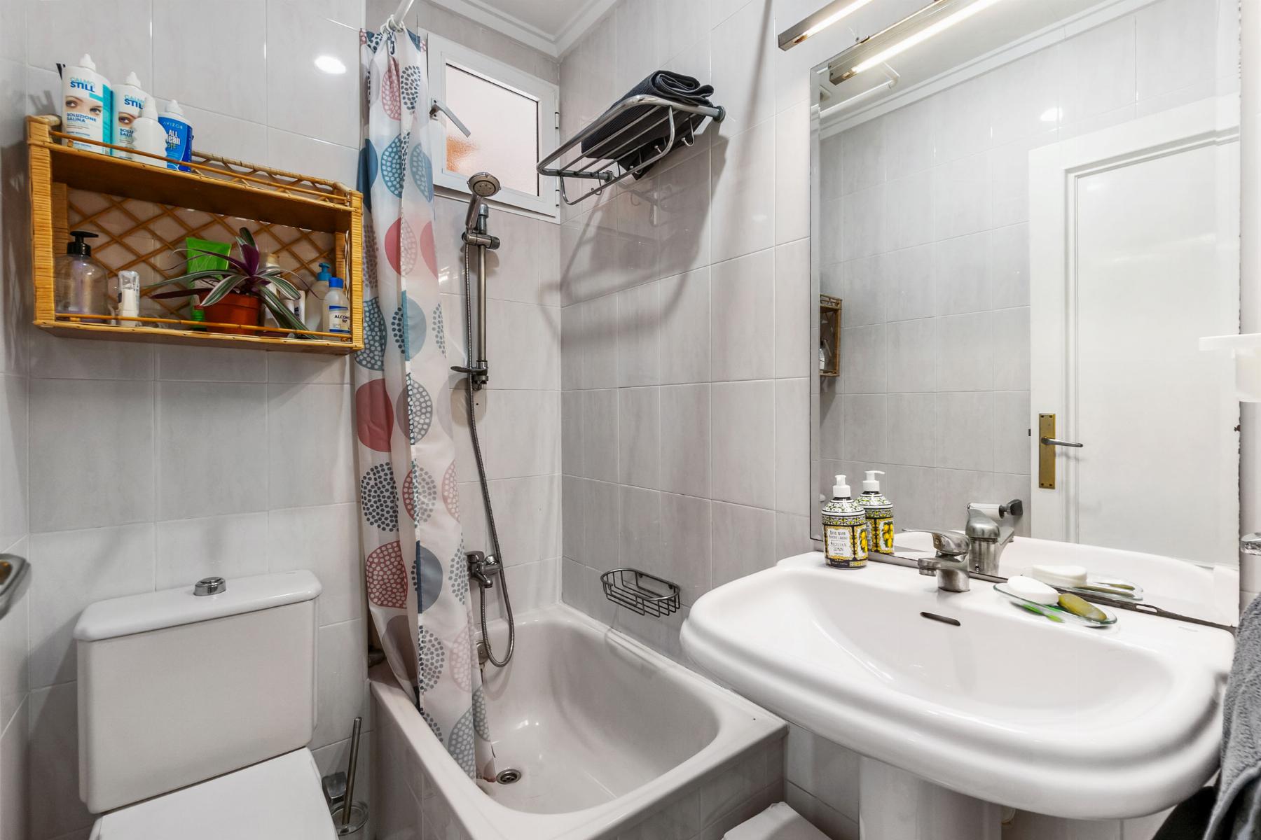 Small white-tiled bathroom with toilet, bathtub/shower combo, colorful patterned curtain, wall shelf of toiletries, and sink with pump bottles.