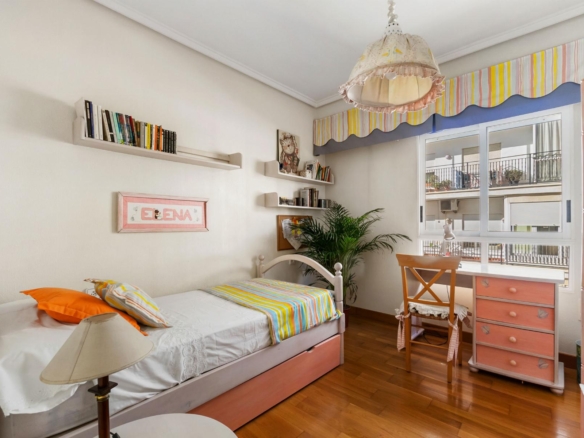 Bright girl's bedroom with white walls, striped rainbow bedding, and a window view of a balcony corner. (informative)