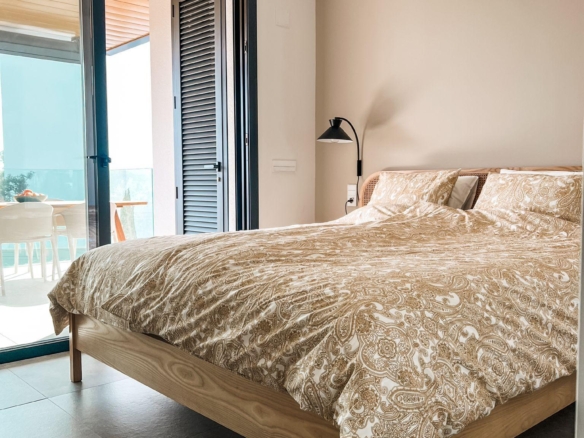 Bedroom with a wooden bed frame and beige paisley bedding; open glass door leads to a balcony with outdoor seating.