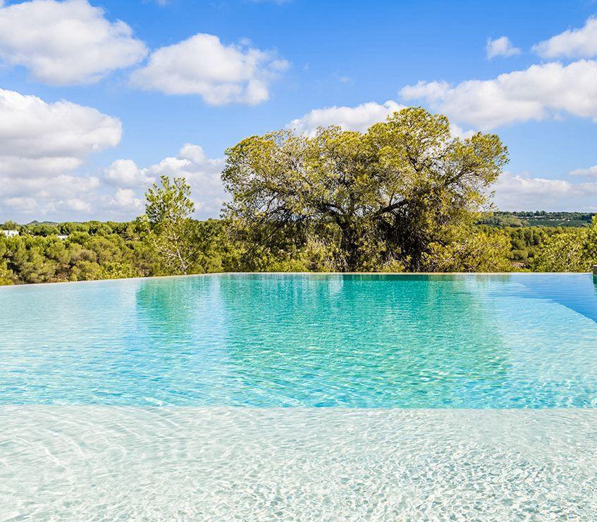 Infinity pool with turquoise water blending into a tree-filled landscape under a blue sky with white clouds