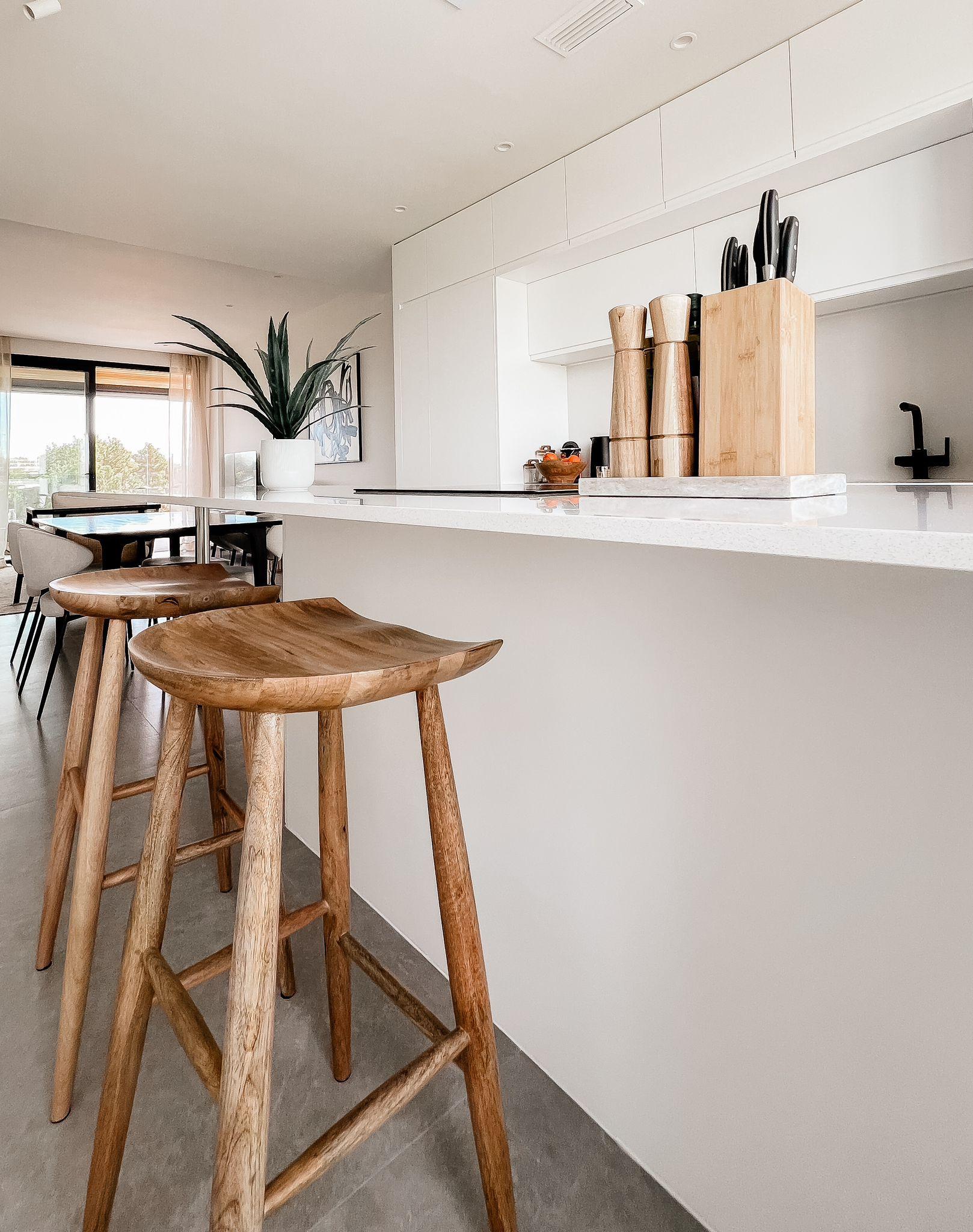 Modern white kitchen with marble countertop, wooden bar stools, and a knife block on the island.