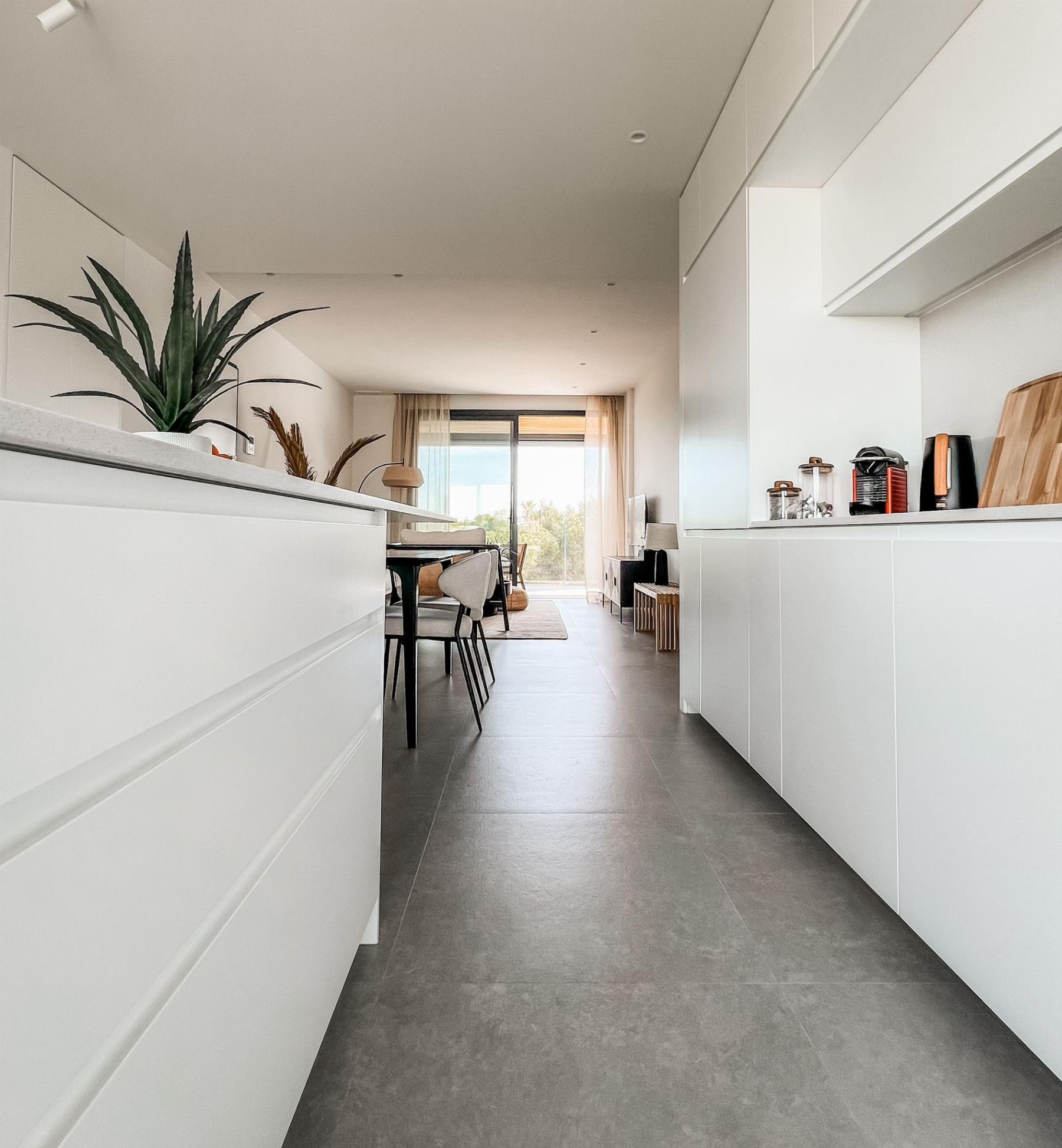 Bright modern kitchen with white glossy cabinets, a long island, and a dining area visible in the distance near a large window.