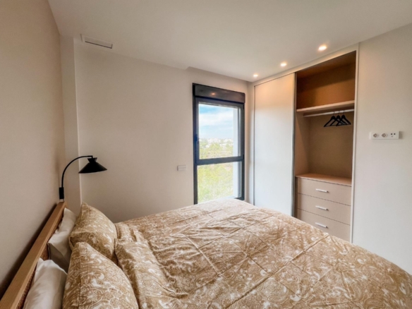 Double bed with beige patterned bedding sits against the left wall, a black reading lamp to the side, and a large window with a city/treetop view nearby beside a built-in wardrobe with hanging space and drawers.