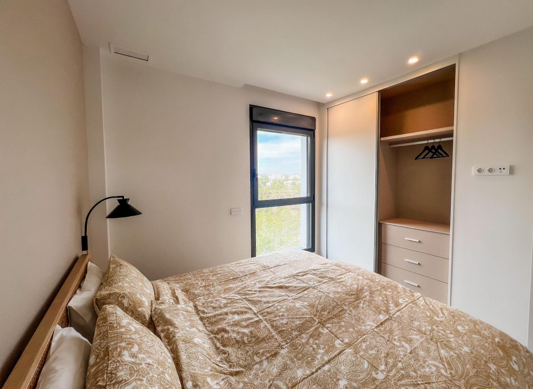 Double bed with beige patterned bedding sits against the left wall, a black reading lamp to the side, and a large window with a city/treetop view nearby beside a built-in wardrobe with hanging space and drawers.