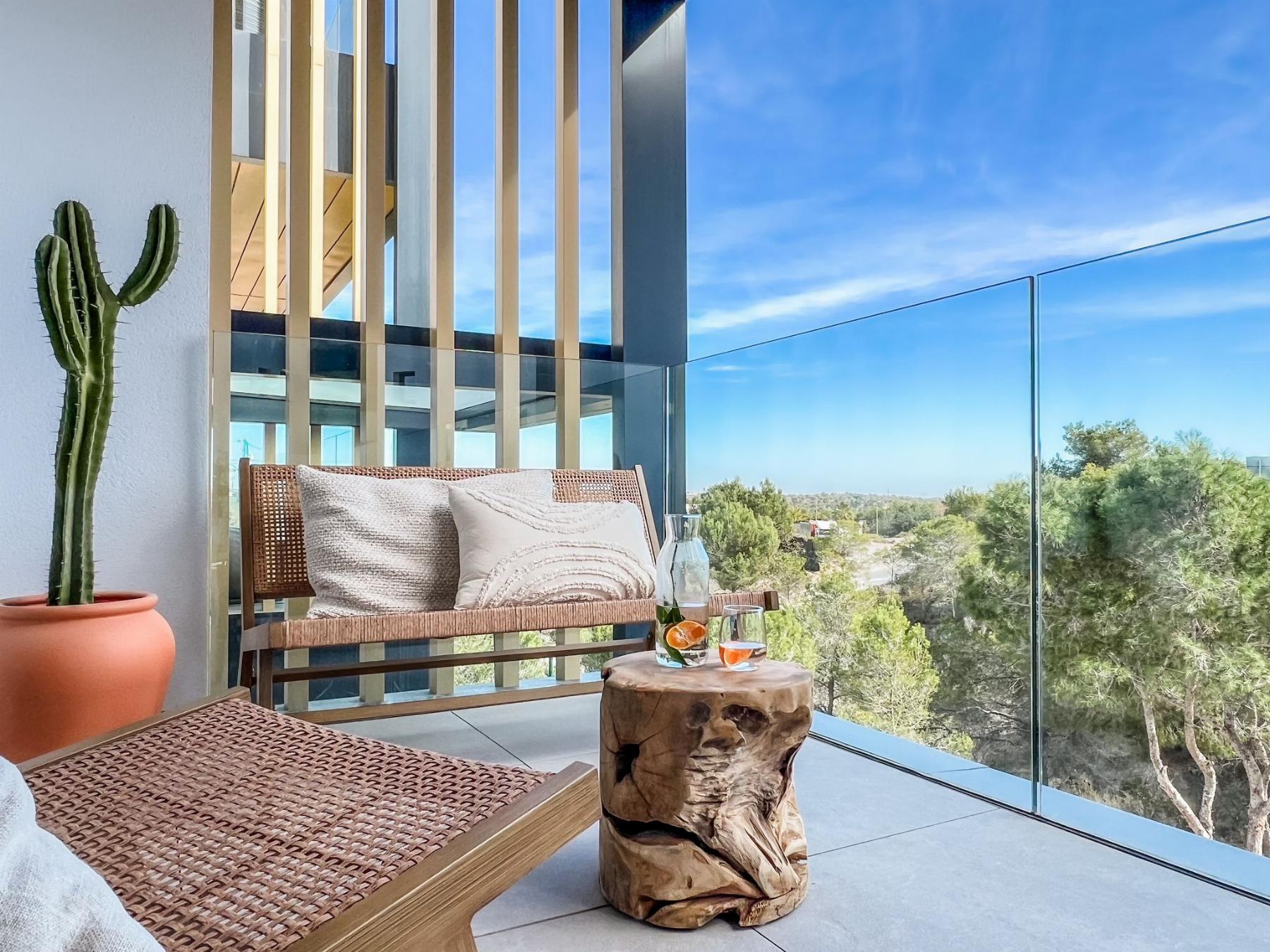 Terracotta pot with a tall cactus beside a wicker lounge chair on a modern balcony, glass railing and blue sky beyond, with cozy beige cushions.