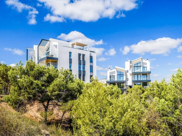 Modern white apartment buildings rise above green shrubs under a bright blue sky with a few clouds in the background.
