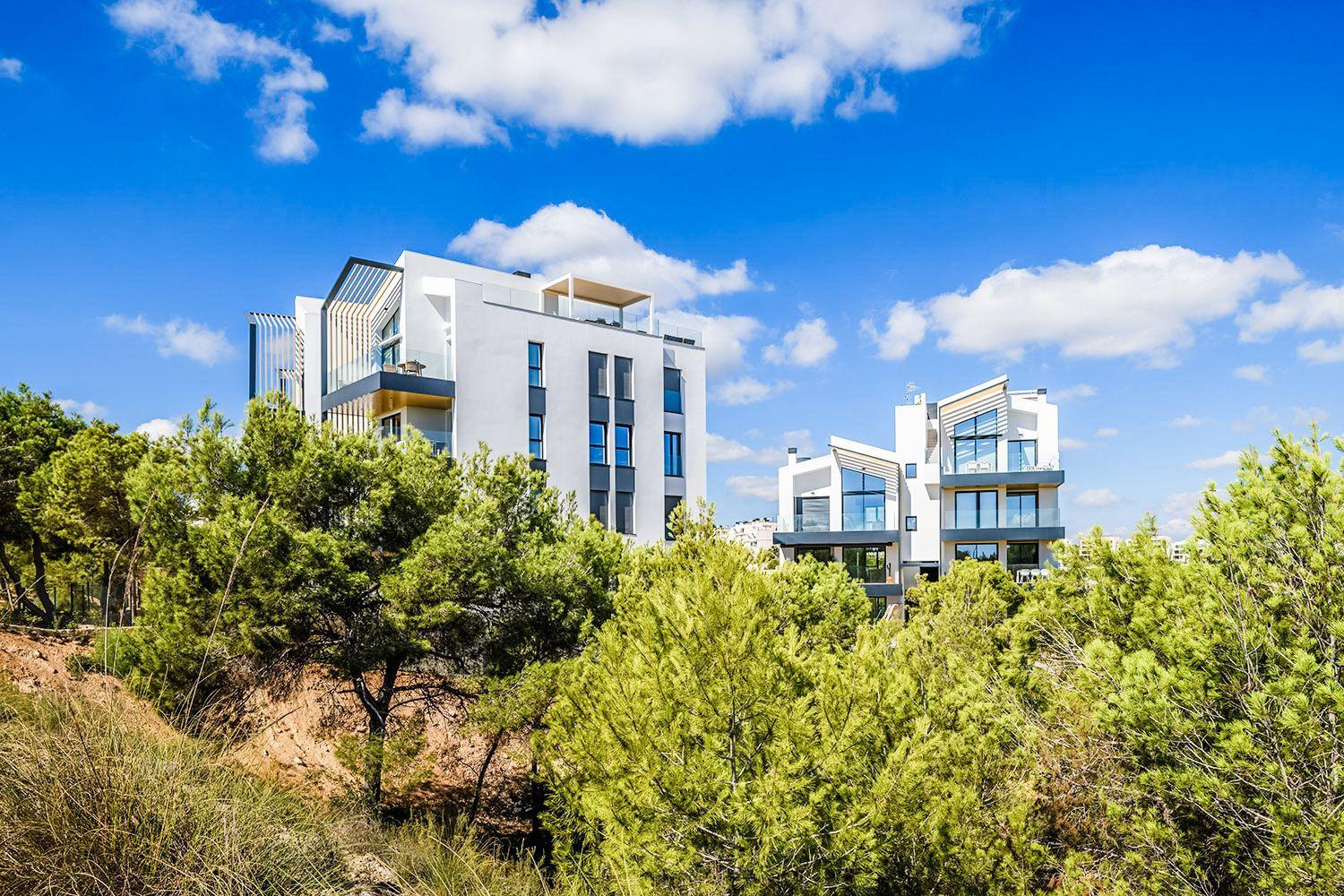 Modern white apartment buildings rise above green shrubs under a bright blue sky with a few clouds in the background.