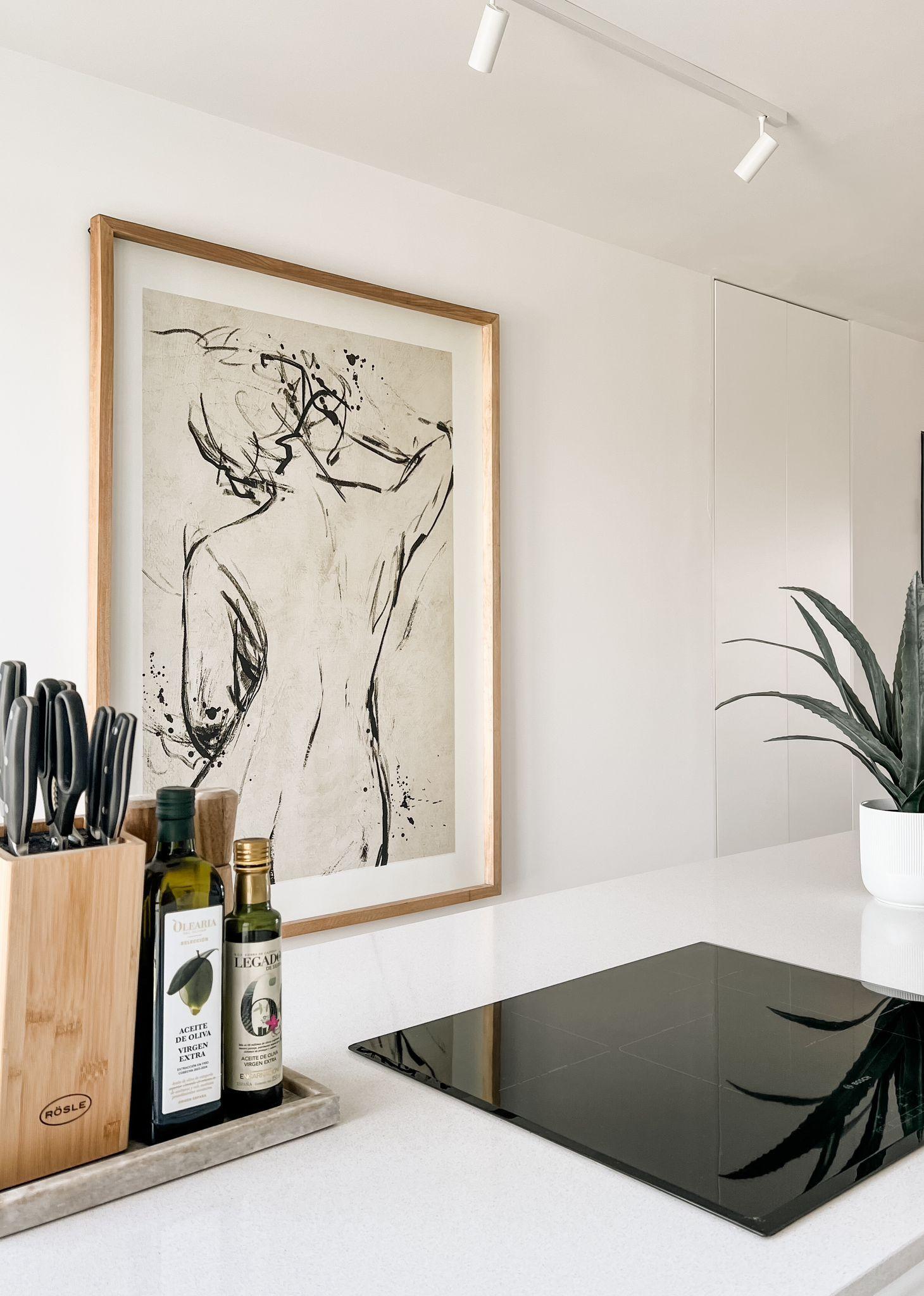 Kitchen counter with a wooden knife block, two bottles of olive oil, and a framed abstract line-art poster leaning against a white wall.