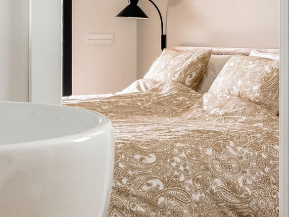 Hotel bedroom scene with a paisley-patterned duvet, pillows, and a black curved wall lamp above the bed.