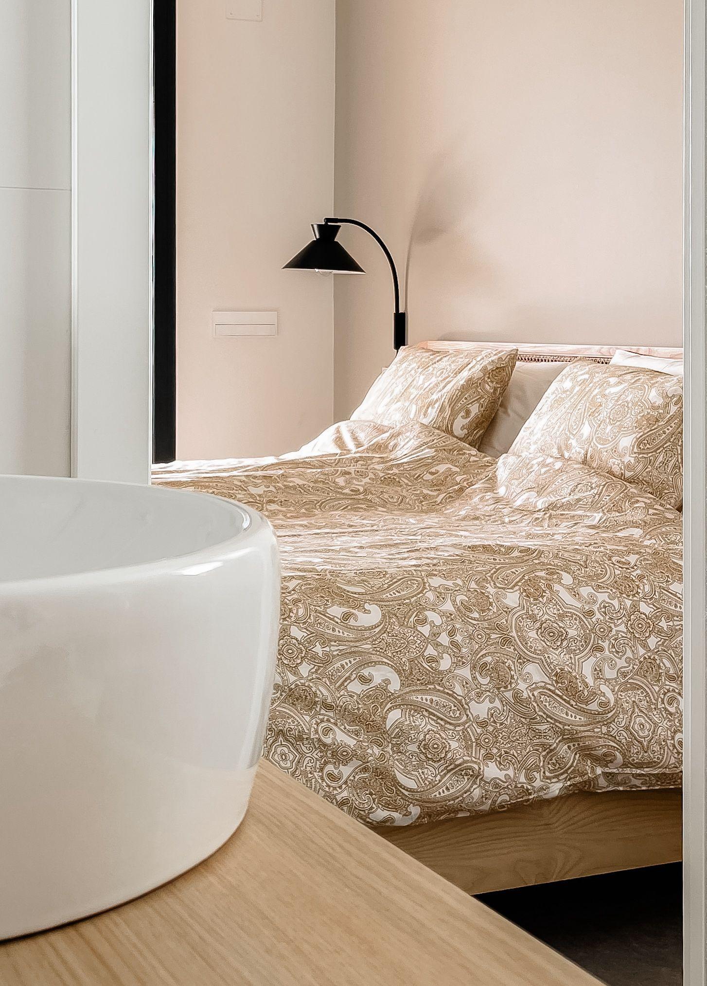 Hotel bedroom scene with a paisley-patterned duvet, pillows, and a black curved wall lamp above the bed.
