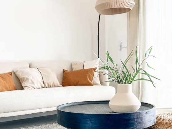 Cozy living room with a cream sofa and orange/beige cushions, a large round blue coffee table, pleated beige floor lamp, and green plants by a window.