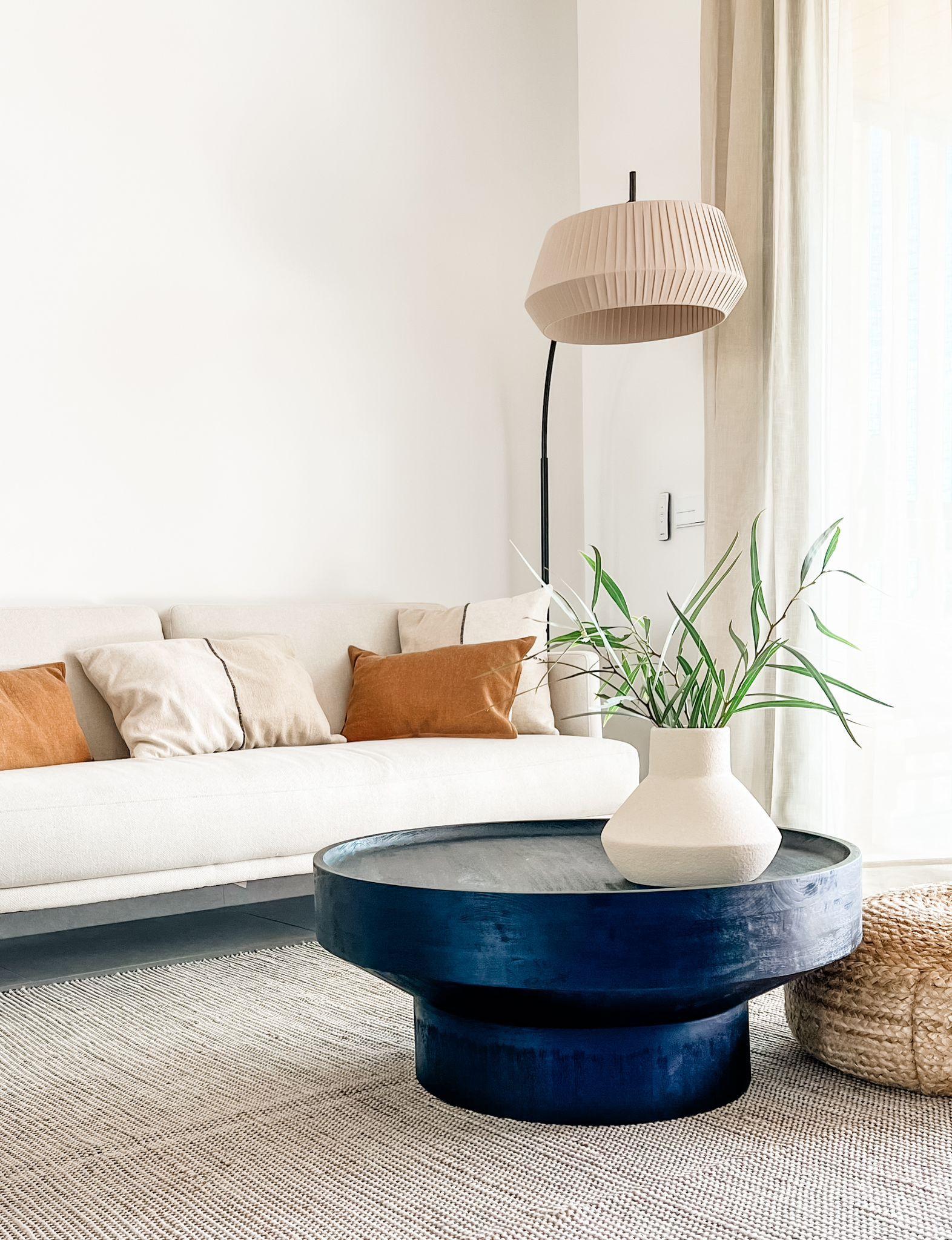 Cozy living room with a cream sofa and orange/beige cushions, a large round blue coffee table, pleated beige floor lamp, and green plants by a window.