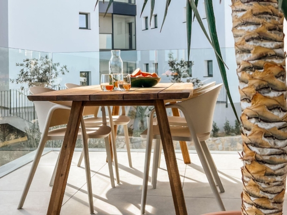 Outdoor dining setup on a sunny balcony: wooden table, white chairs, glass railing, and a palm tree in the foreground.
