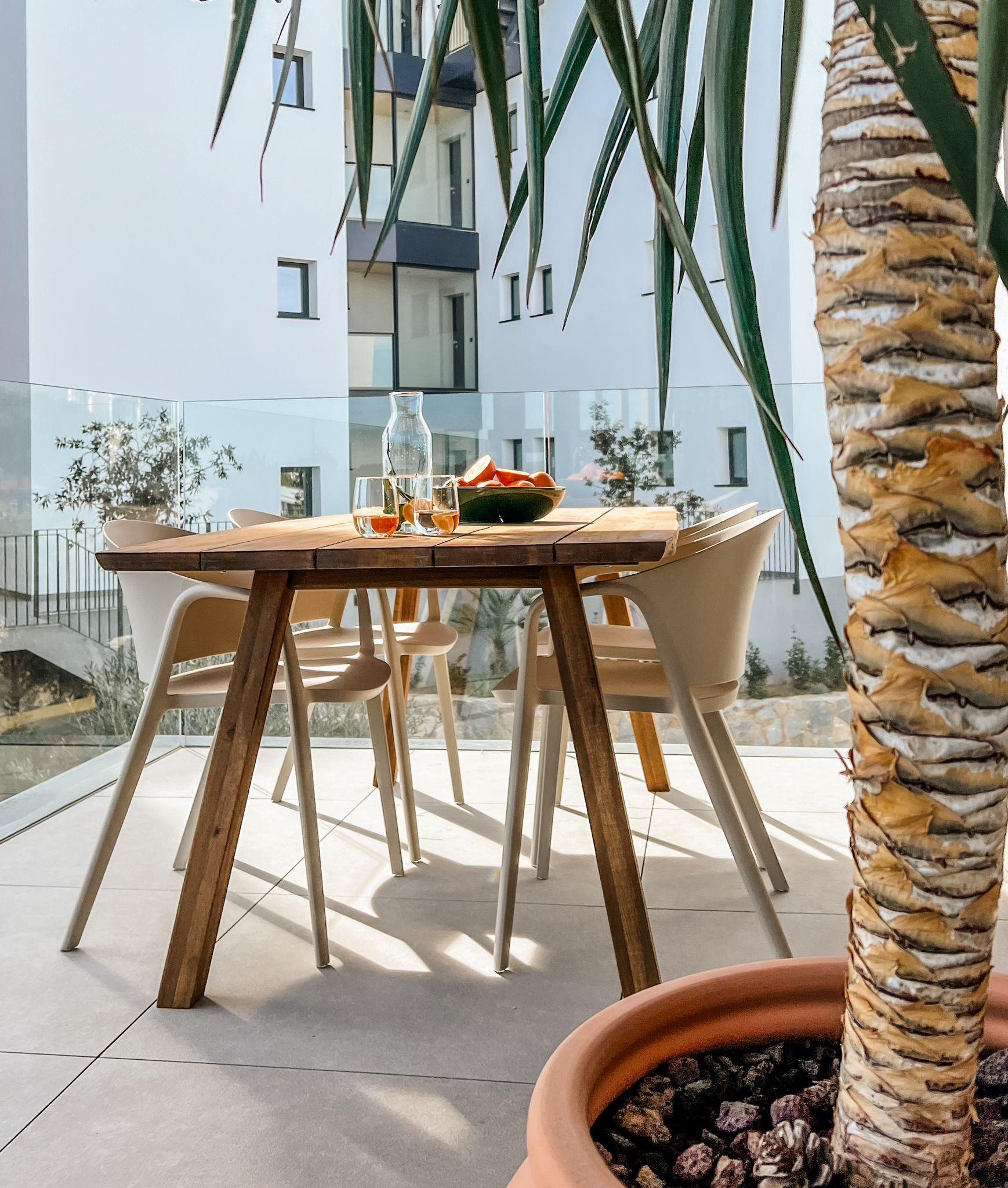 Outdoor dining setup on a sunny balcony: wooden table, white chairs, glass railing, and a palm tree in the foreground.