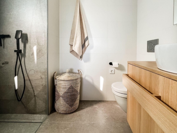 Modern bathroom with a glass shower on the left, beige towel on a wall hook, a woven basket, and a wooden vanity with a white basin on the right.