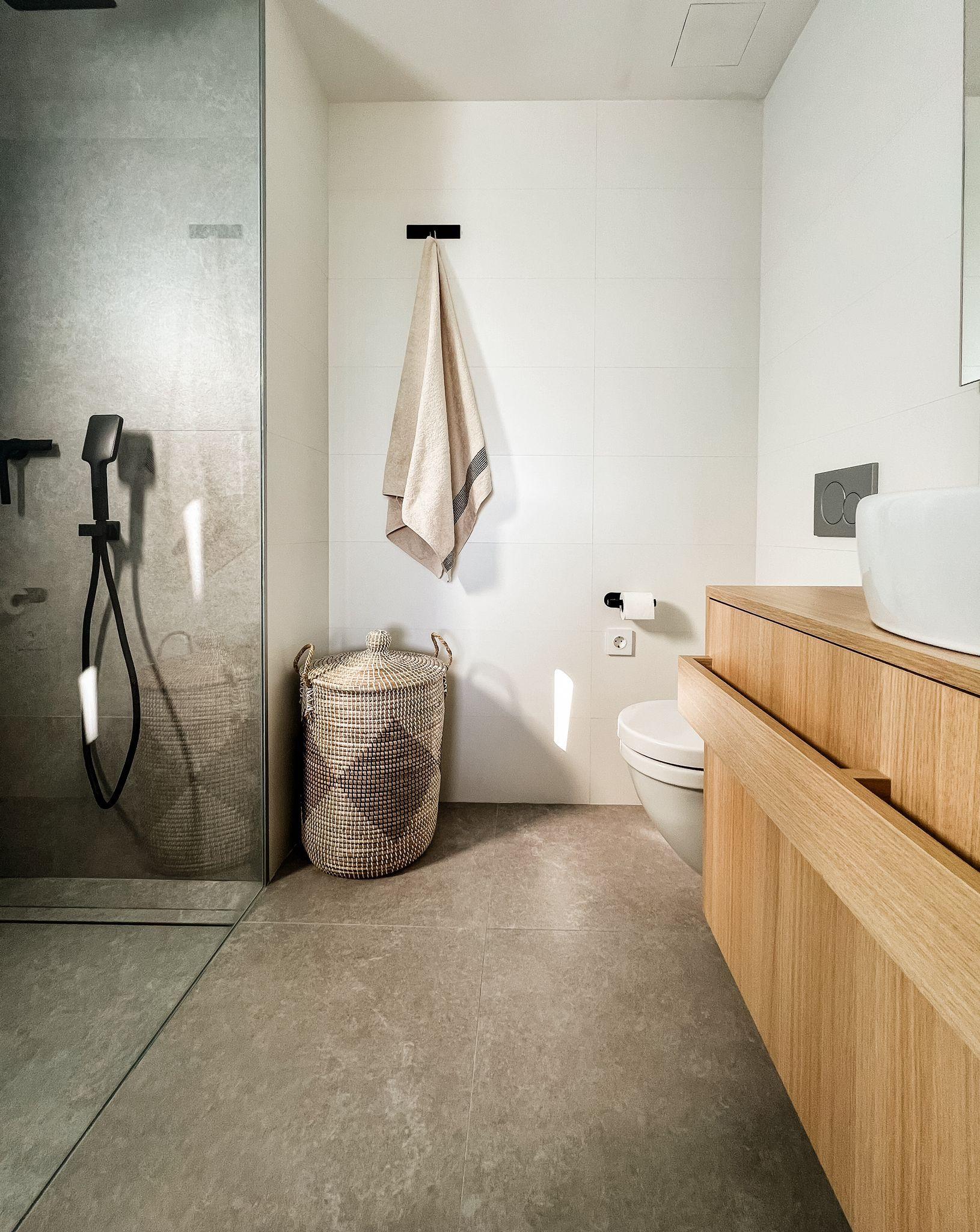Modern bathroom with a glass shower on the left, beige towel on a wall hook, a woven basket, and a wooden vanity with a white basin on the right.