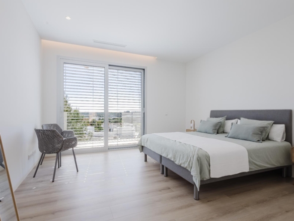 Bright, modern bedroom with a gray upholstered bed, pale green pillows, and white bedding near a large sliding glass door with horizontal blinds. The view shows trees outside.