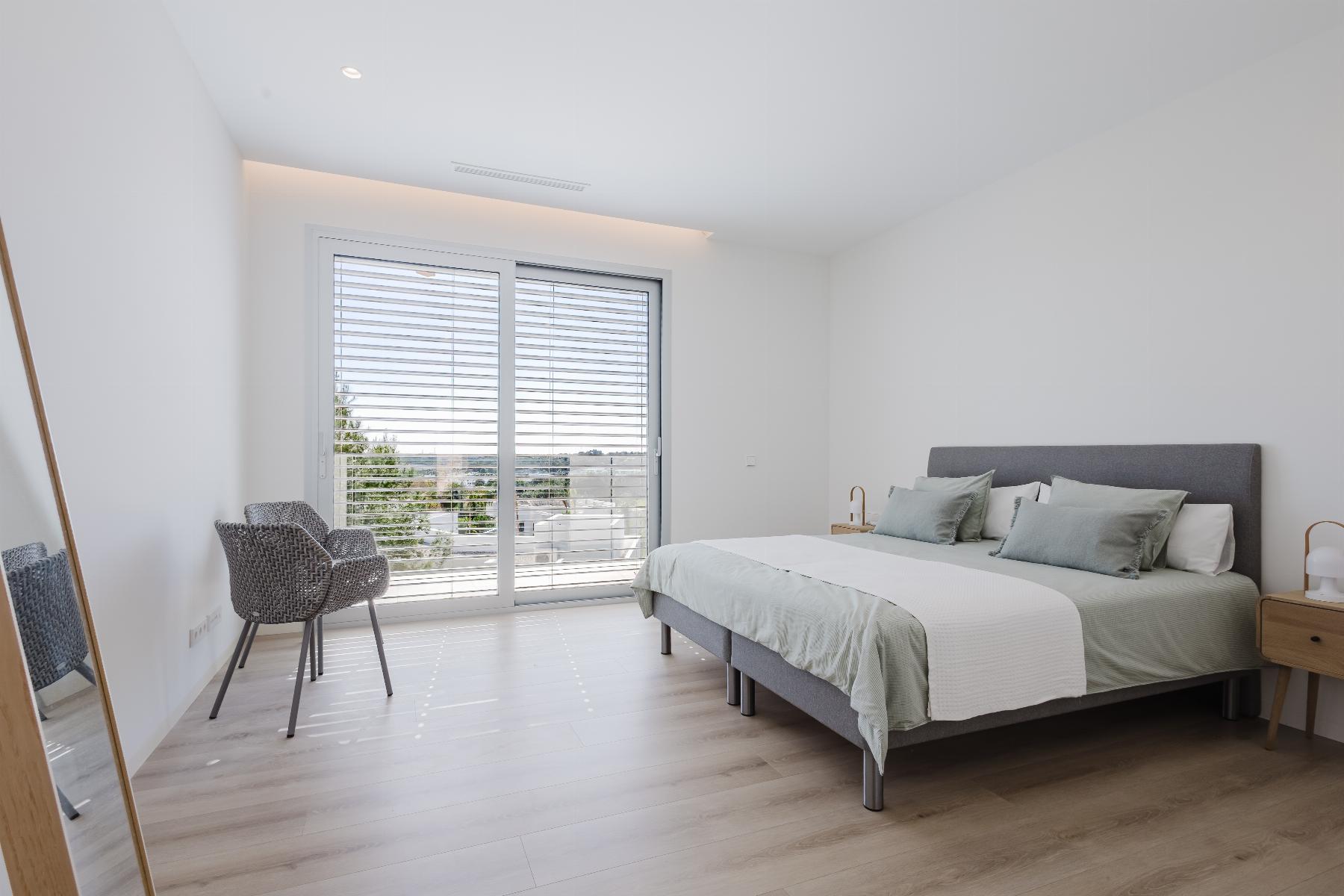 Bright, modern bedroom with a gray upholstered bed, pale green pillows, and white bedding near a large sliding glass door with horizontal blinds. The view shows trees outside.