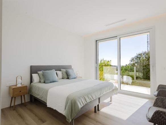 Bright modern bedroom with a gray upholstered bed, pale green and white bedding, and two small wooden nightstands with lamps; large sliding glass doors open to a balcony with greenery outside.