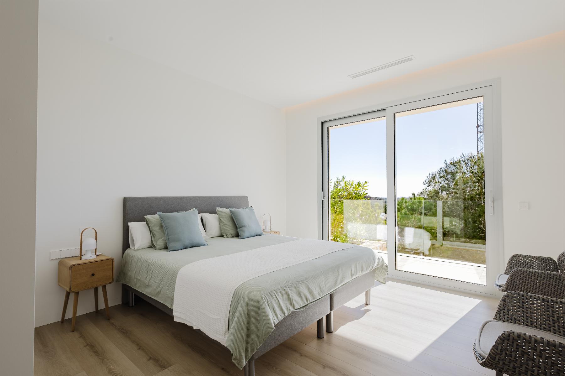 Bright modern bedroom with a gray upholstered bed, pale green and white bedding, and two small wooden nightstands with lamps; large sliding glass doors open to a balcony with greenery outside.