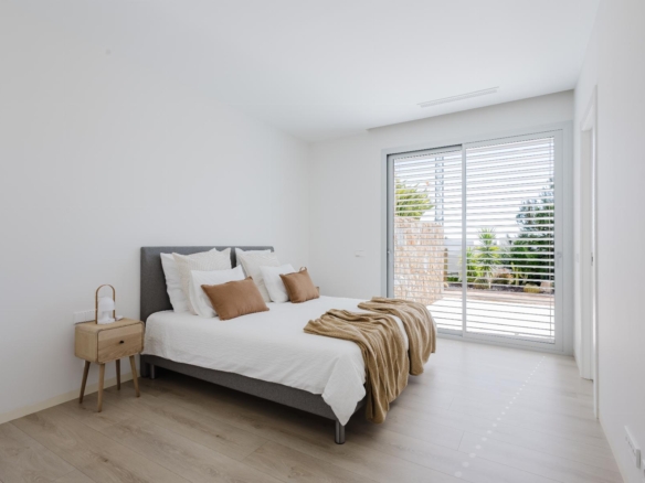 Bright minimalist bedroom with white walls, a gray upholstered bed, white bedding, and tan accent pillows with a wooden nightstand nearby. a blinds-sliding door to the outside.