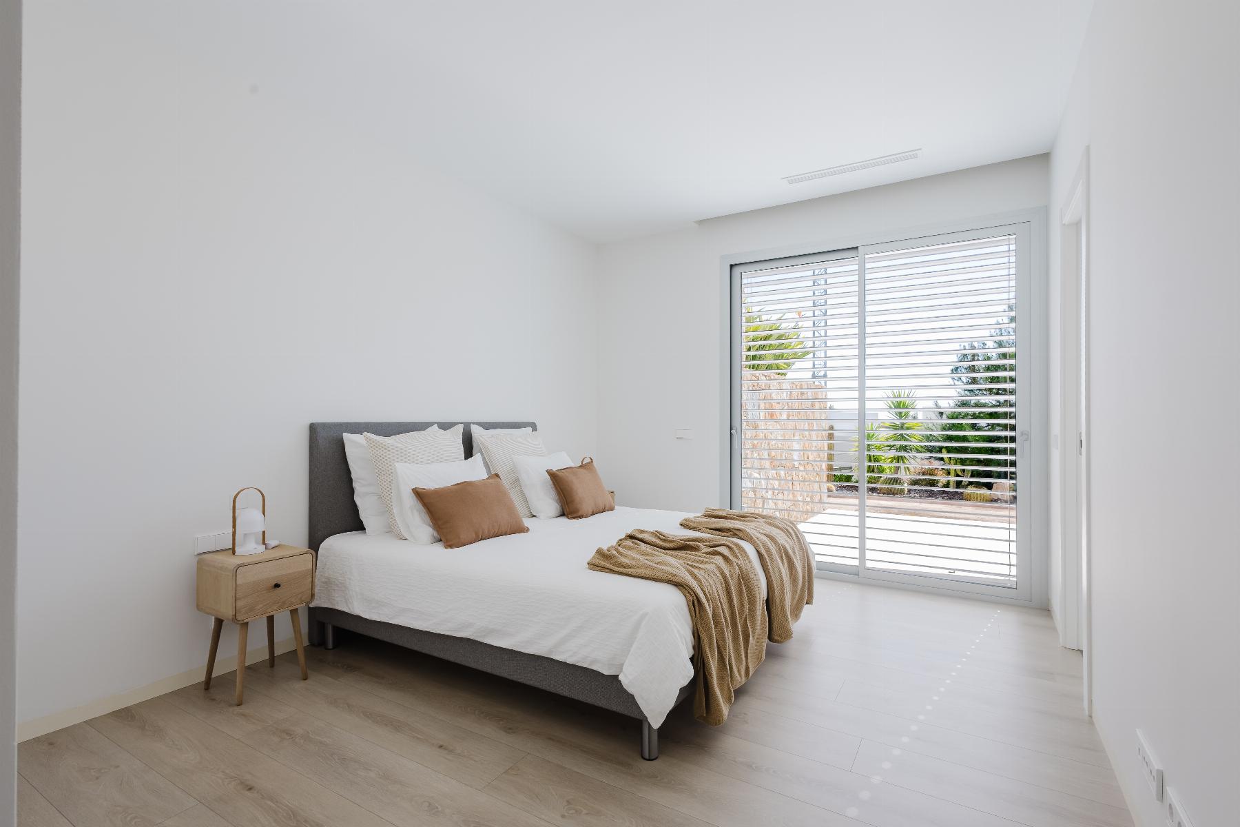 Bright minimalist bedroom with white walls, a gray upholstered bed, white bedding, and tan accent pillows with a wooden nightstand nearby. a blinds-sliding door to the outside.