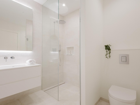 Minimalist white bathroom with frameless glass shower, floating vanity, and wall-mounted toilet; small plant on a shelf nearby.