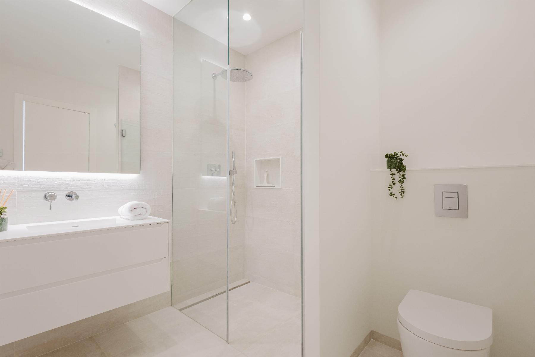 Minimalist white bathroom with frameless glass shower, floating vanity, and wall-mounted toilet; small plant on a shelf nearby.