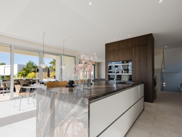 Modern open-plan kitchen with a marble island, dark wood cabinetry, and built-in ovens; sliding glass doors open to an outdoor dining area