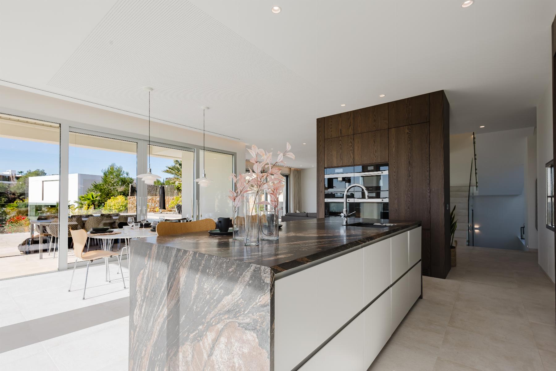 Modern open-plan kitchen with a marble island, dark wood cabinetry, and built-in ovens; sliding glass doors open to an outdoor dining area