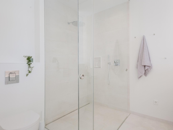 Minimalist white bathroom featuring a glass-walled shower, chrome fixtures, and a small plant on the wall shelf.