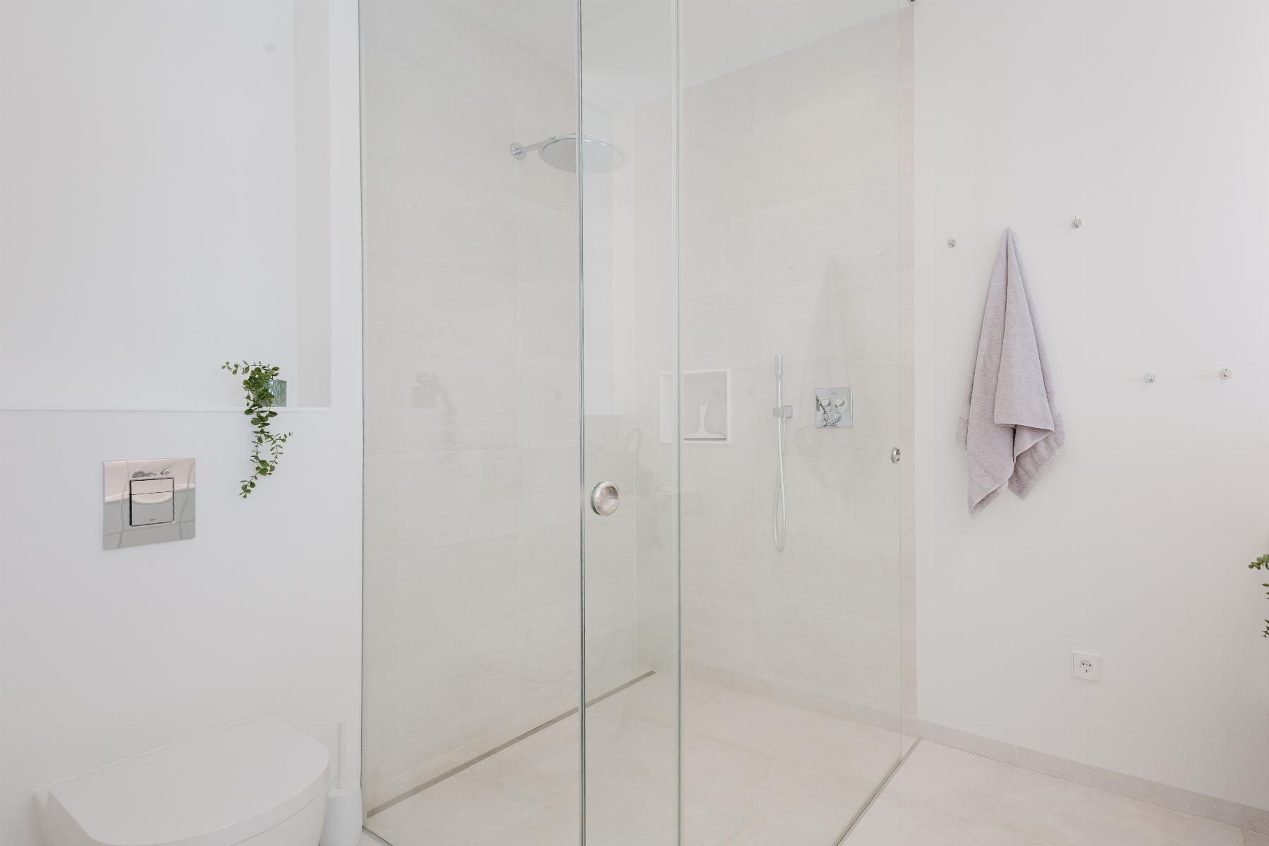 Minimalist white bathroom featuring a glass-walled shower, chrome fixtures, and a small plant on the wall shelf.