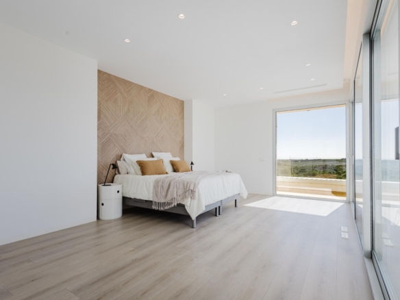 Bright, minimalist bedroom with a low platform bed, woven headboard, white bedding, and neutral pillows; large glass doors open to a balcony with a landscape view.