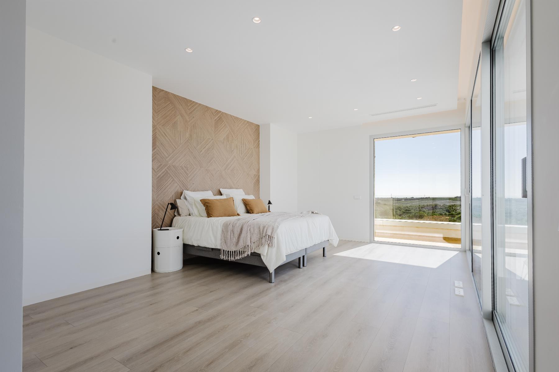 Bright, minimalist bedroom with a low platform bed, woven headboard, white bedding, and neutral pillows; large glass doors open to a balcony with a landscape view.
